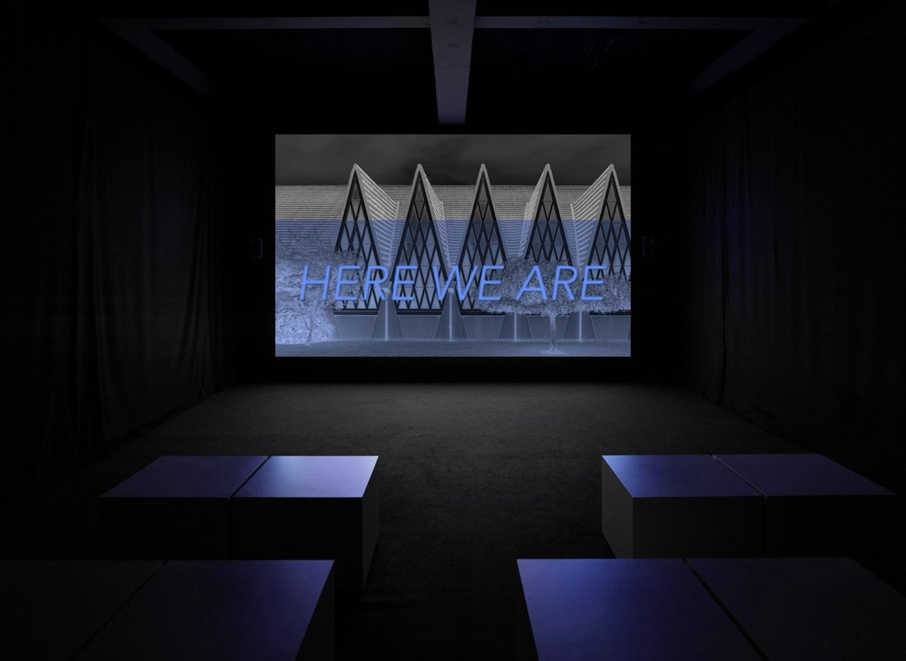 A darkened gallery with black drapes. A projection shows a row of tall tent-like white structures behind a wall, with the words HERE WE ARE across them in blue.
