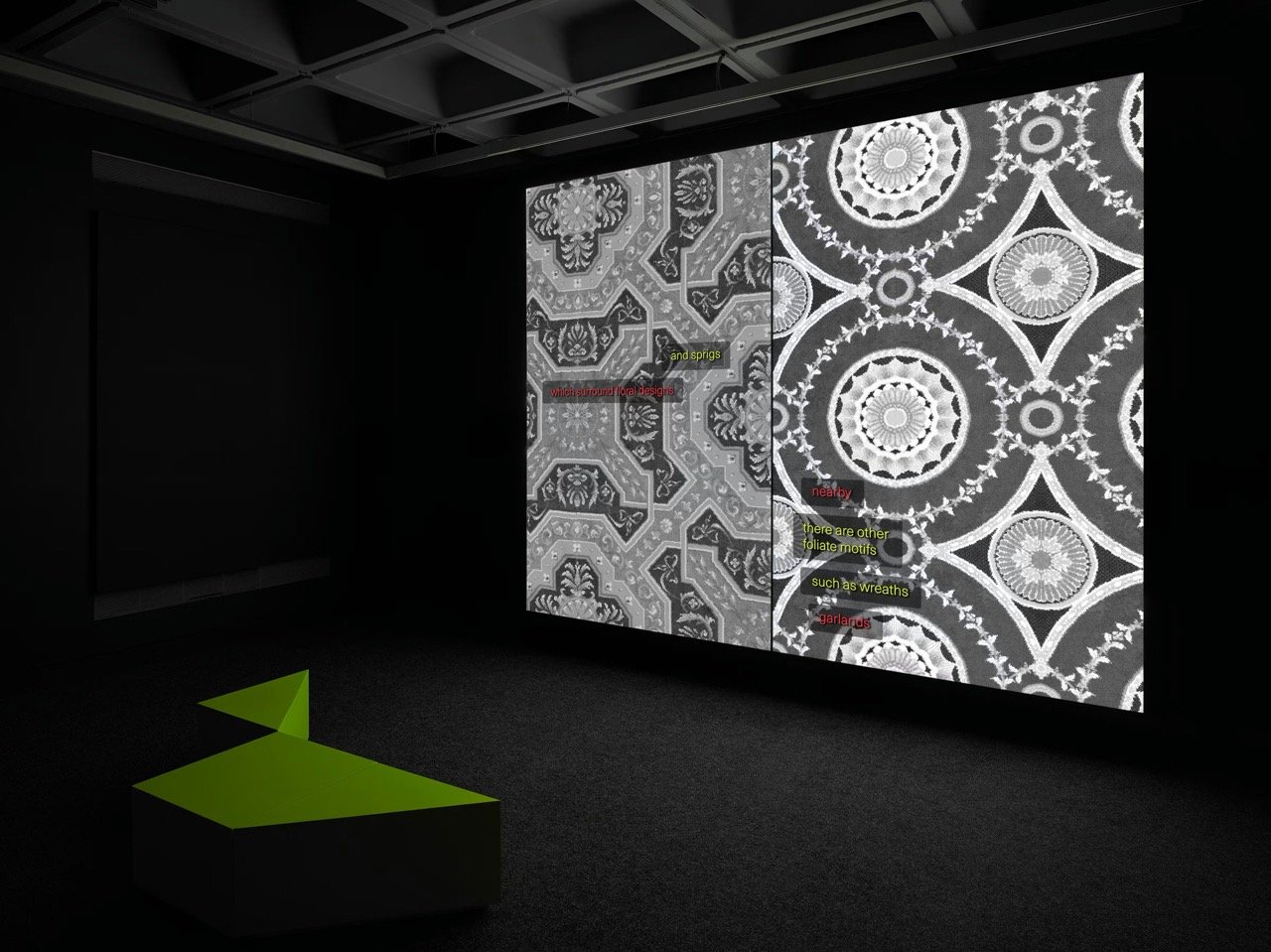 A dark gallery with a coffered ceiling. A large projection fills the wall with two ornate grey carpet patterns side by side, small lines of coloured text overlaid on them. An angular green bench sits on the dark carpet in front.