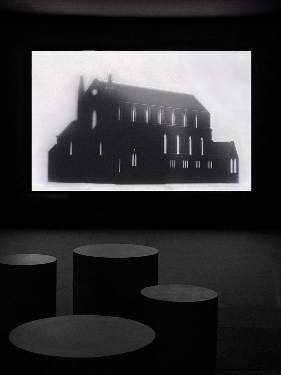 A darkened gallery. A projection shows a church in negative, its silhouette black against a white sky, windows glowing as pale slots. Low cylindrical seating stands on the floor in front of the screen.