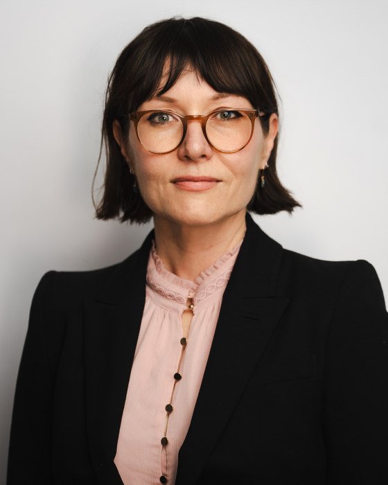 Leah Kardos in a black blazer and pale pink blouse against a light grey studio backdrop, 2026