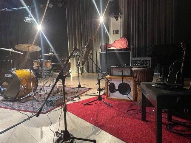 A live room set up for recording, with microphones, a guitar amplifier and a drum kit on a red rug.